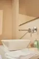 Wand-Waschtischarmatur in Chrom, montiert an der Wand über einem weißen, quadratischen Waschbecken. Ideal Standard-Design mit Handtuch und Pflegeprodukten im Hintergrund.